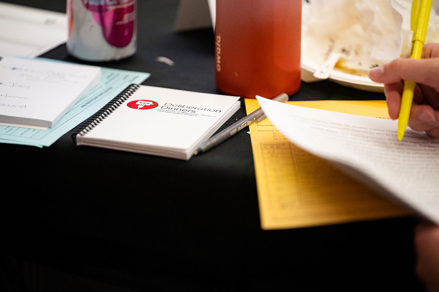 A notepad with the Deliberation Dinner logo on it is on a table with other papers, with a close up of a hand that is using a highlighter.